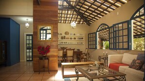 Un salon spacieux avec un plafond haut, des poutres apparentes, une table basse en verre, un canapé blanc et un coin repas avec des meubles en bois.