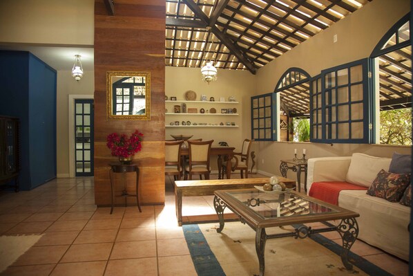 A spacious living room with a high ceiling, wooden beams, a glass coffee table, a white sofa, and a dining area with wooden furniture.
