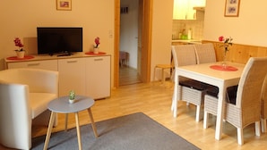 A modern living room with a flat-screen TV, a dining area with wicker chairs, and a kitchen visible through an open door. - Vacation apartment in Hochkönig, hiking possibilities just in front of the door (Mühlbach/Hochkönig)