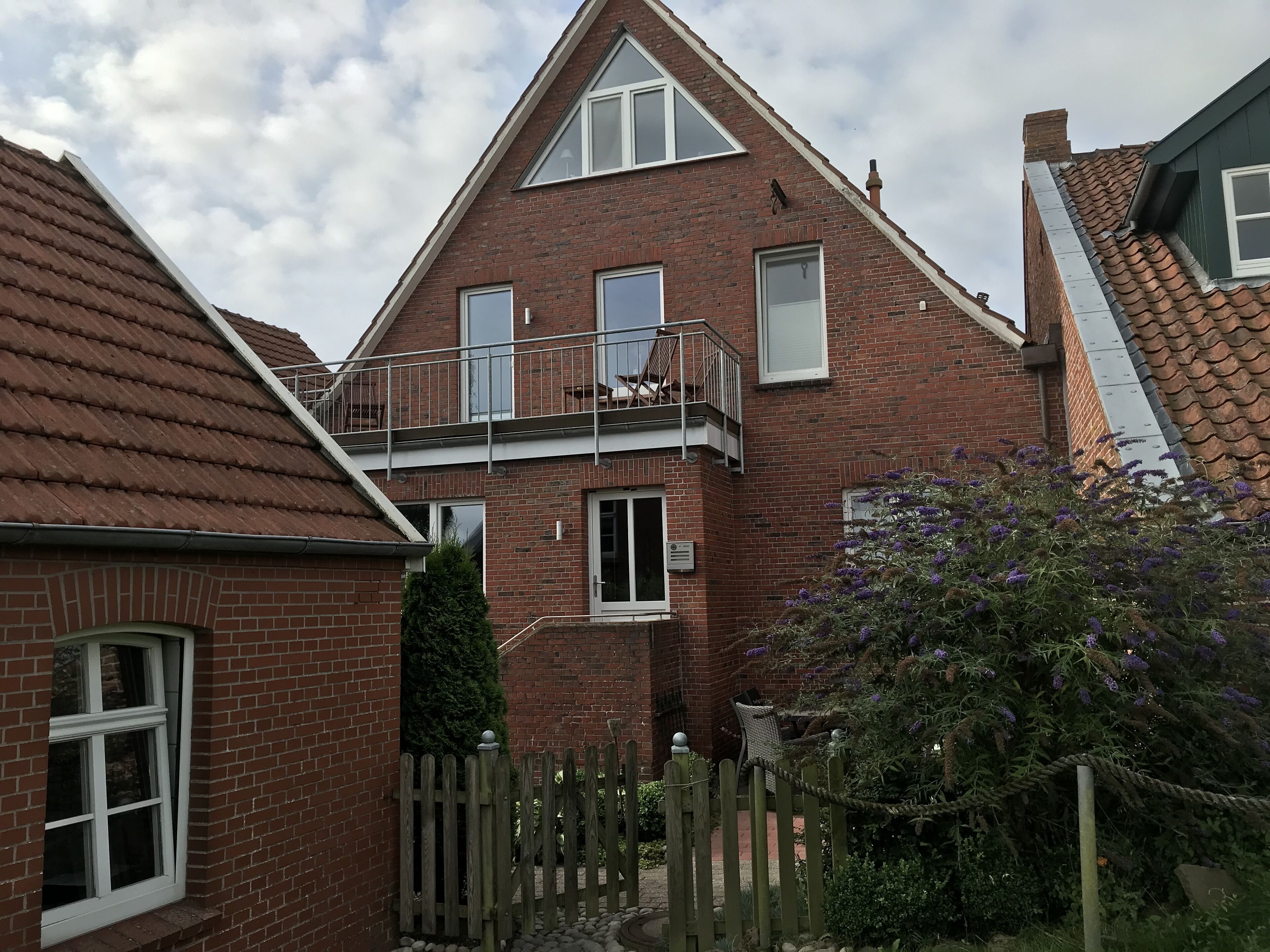 A two-story brick house with a balcony, a wooden fence, and a garden with purple flowers.