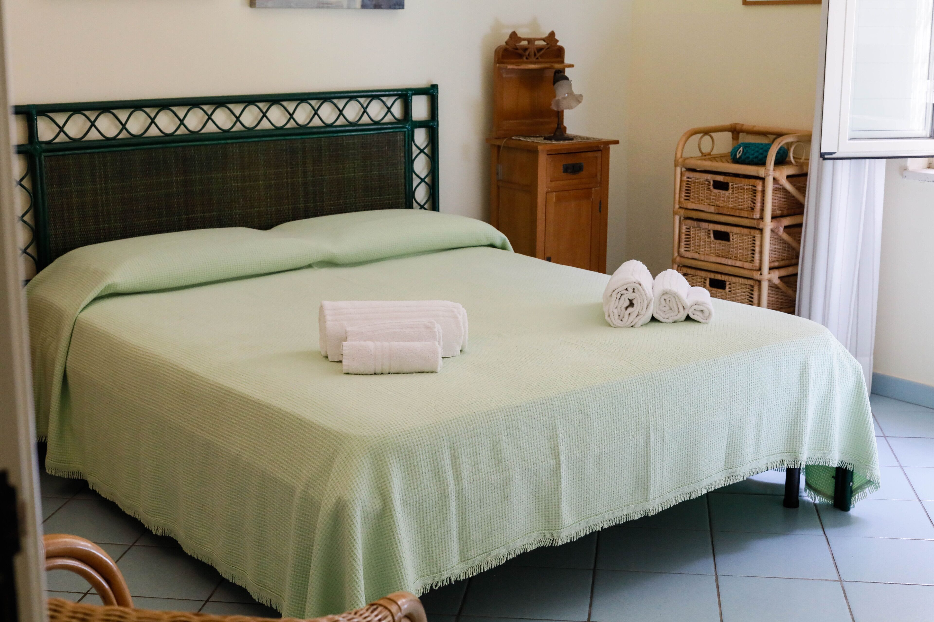 A neatly made bed with a green bedspread and two rolled towels on the side.