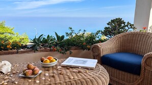 Une chaise en osier avec un coussin, un saladier rempli de fruits et un livre posés sur une table en fibres naturelles, le tout avec vue sur la côte.