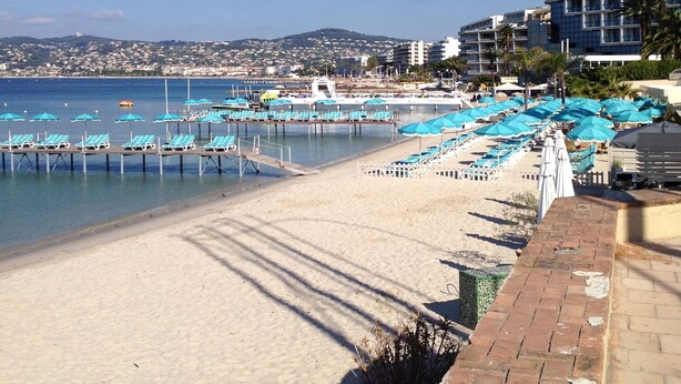 Ein Strand mit Liegestühlen und Sonnenschirmen, einer Pier und Gebäuden im Hintergrund.