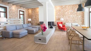 A modern living room with a stone wall, a grey sectional sofa, a red chair, a long dining table with chairs, and a fireplace. - Ferrer de Shorta Modern holiday home for 10, private pool, A/C and Wi-Fi (S'Horta)