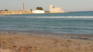 Una spiaggia con un litorale sabbioso, qualche roccia sparsa e un complesso di edifici storici con rovine e un'alta struttura.