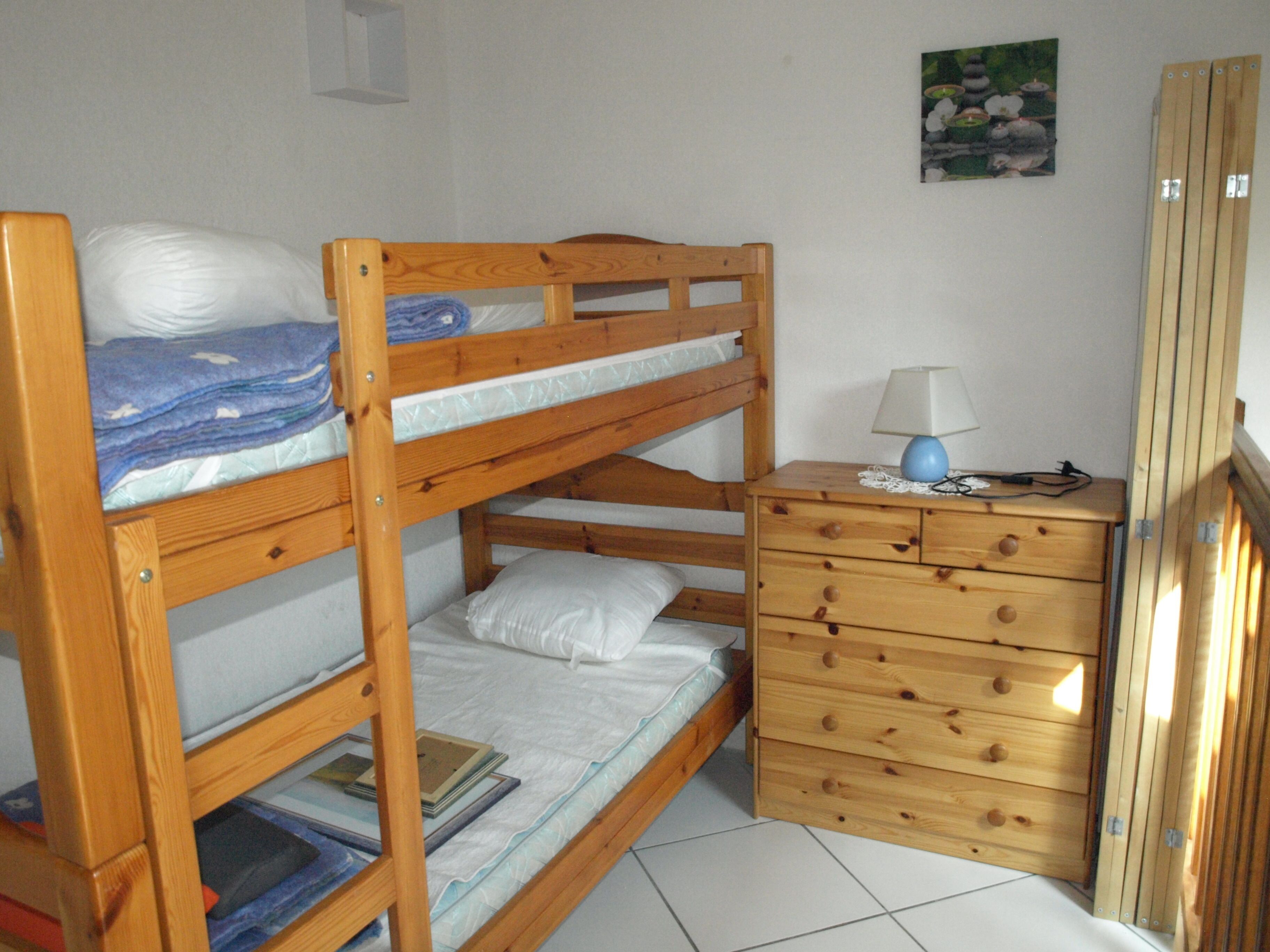Een stapelbed met een houten dressoir ernaast, een klein tafeltje met een lamp, en een ingelijst fotoprint op de muur.