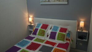 Een net opgemaakt bed met kleurrijke geometrische kussens, twee bedlampen en een schilderij boven het bed.