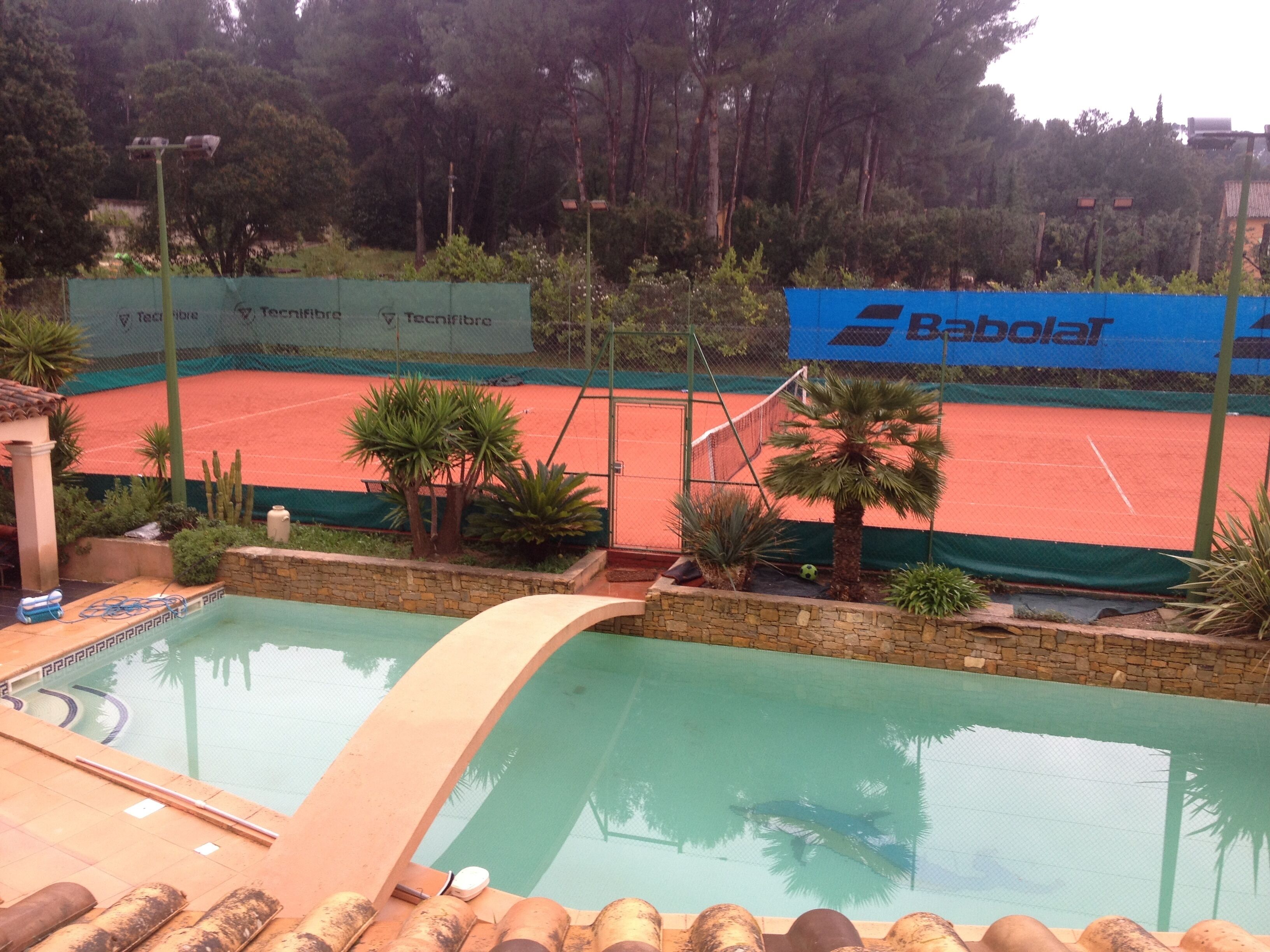 A swimming pool with a tennis court in the background, surrounded by palm trees and a stone wall.