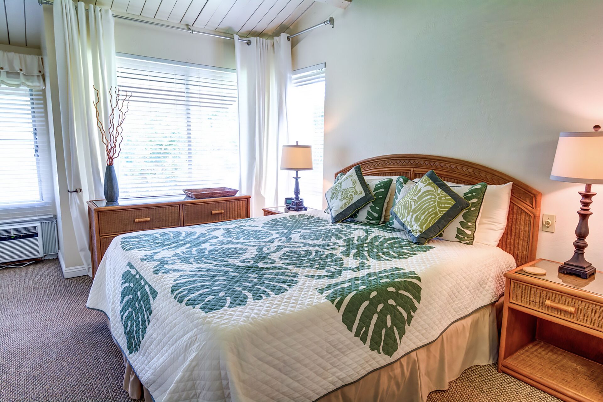 A neatly made bed with a leaf-patterned bedspread, a wooden headboard, and a bedside table with a lamp.