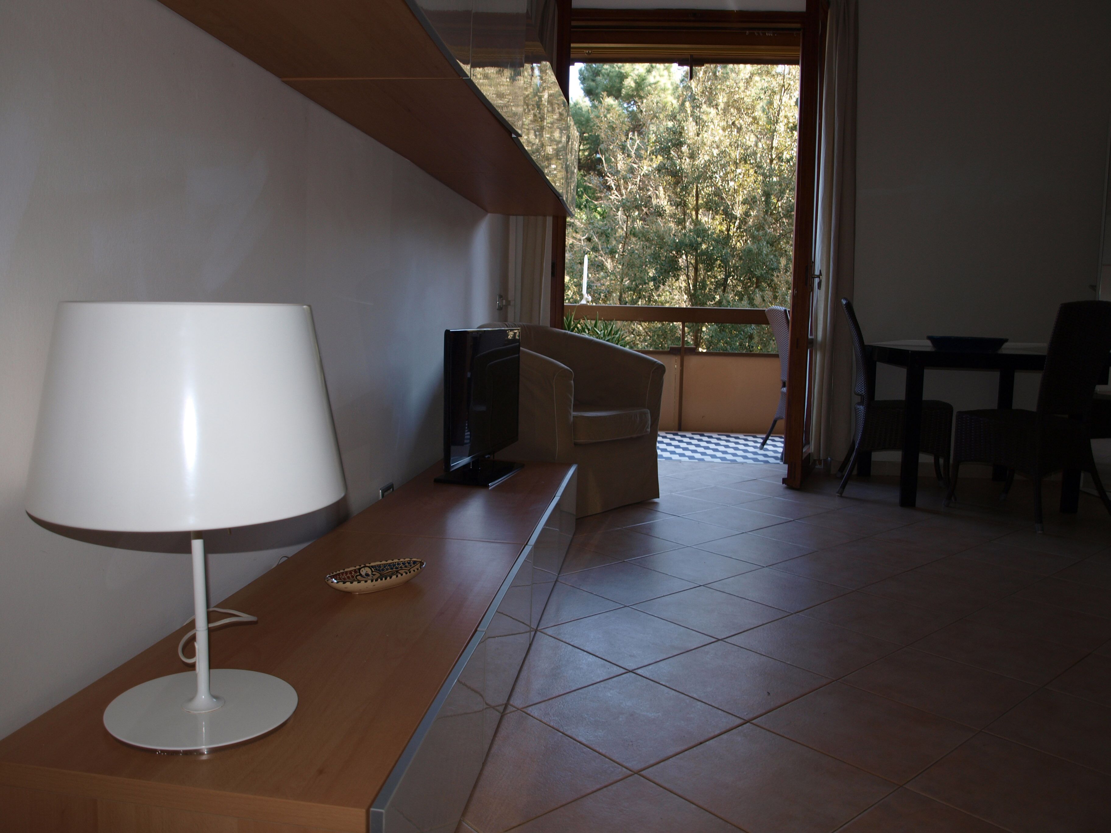 A room with a wooden desk, a white lamp, a television, and a view of trees through a window.