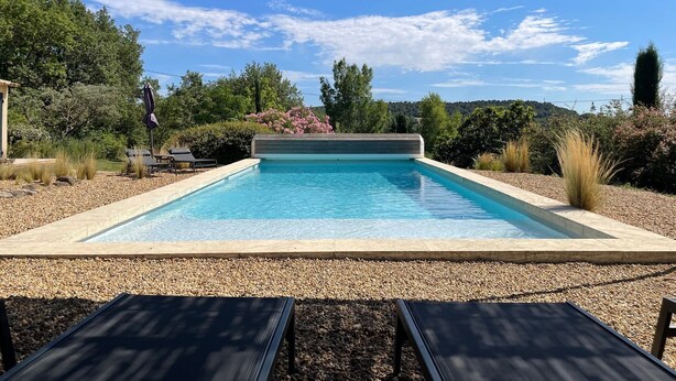 A rectangular swimming pool with clear blue water, surrounded by pebbles and shaded by a pergola.