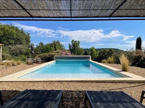 A rectangular swimming pool with clear blue water, surrounded by pebbles and shaded by a pergola.