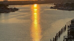 Un coucher de soleil sur une rivière paisible, avec des bateaux amarrés sur la rive droite, une jetée s’avançant dans l’eau et, au loin, le profil d’une ville.