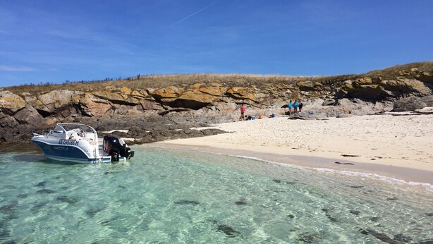 Un bateau est amarré près d’une plage rocheuse, où l’eau est claire et le rivage sablonneux. On aperçoit un groupe de personnes au loin.