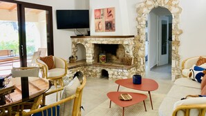 A cozy living room with a stone fireplace, wicker furniture, and a flat-screen TV mounted on the wall. - VILLA en Pierre 4 persons near PORTO-VECCHIO HEATED SWIMMING POOL  (Lecci)
