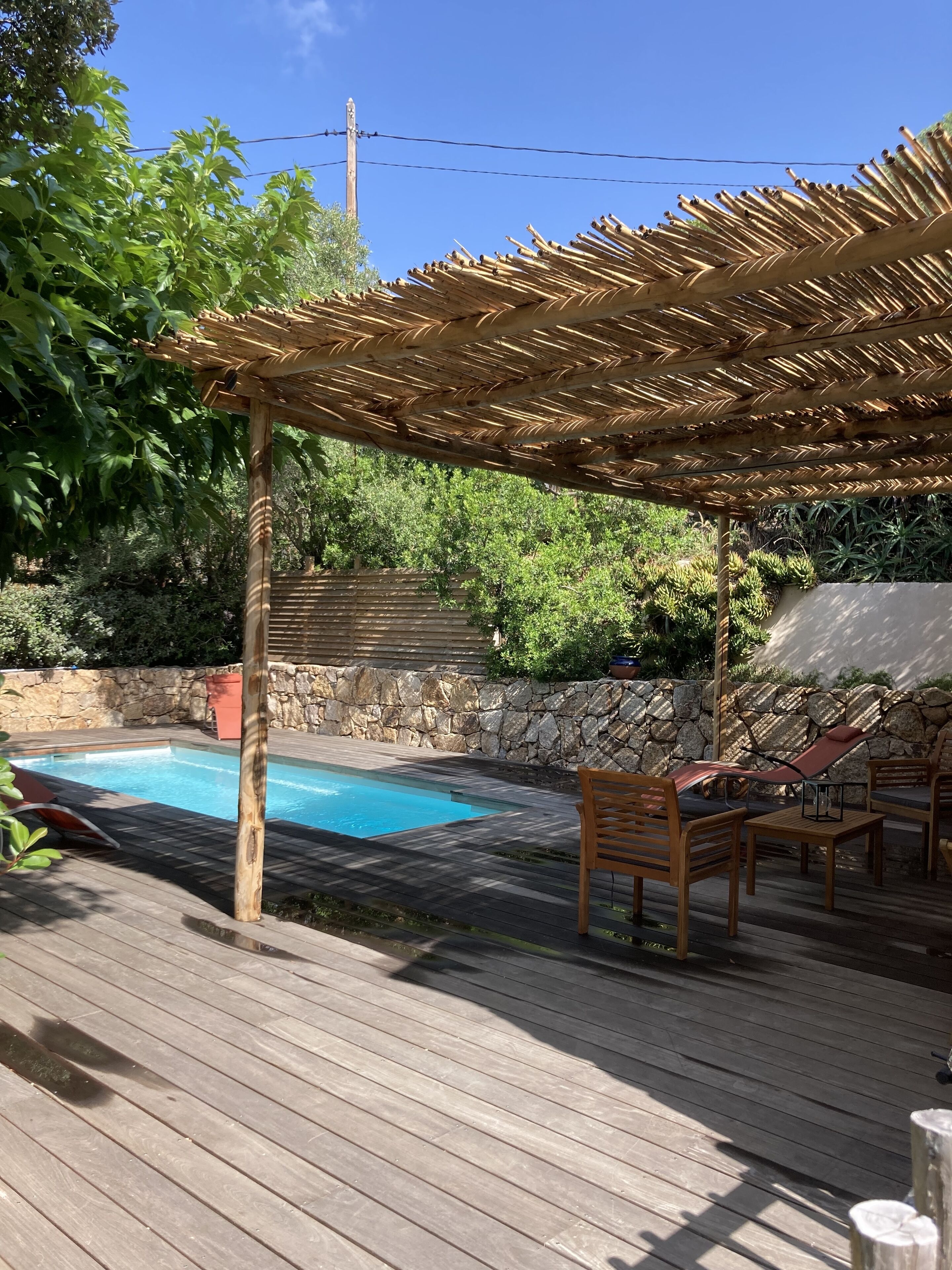 A wooden deck with a pool, stone wall, and a pergola with a thatched roof.