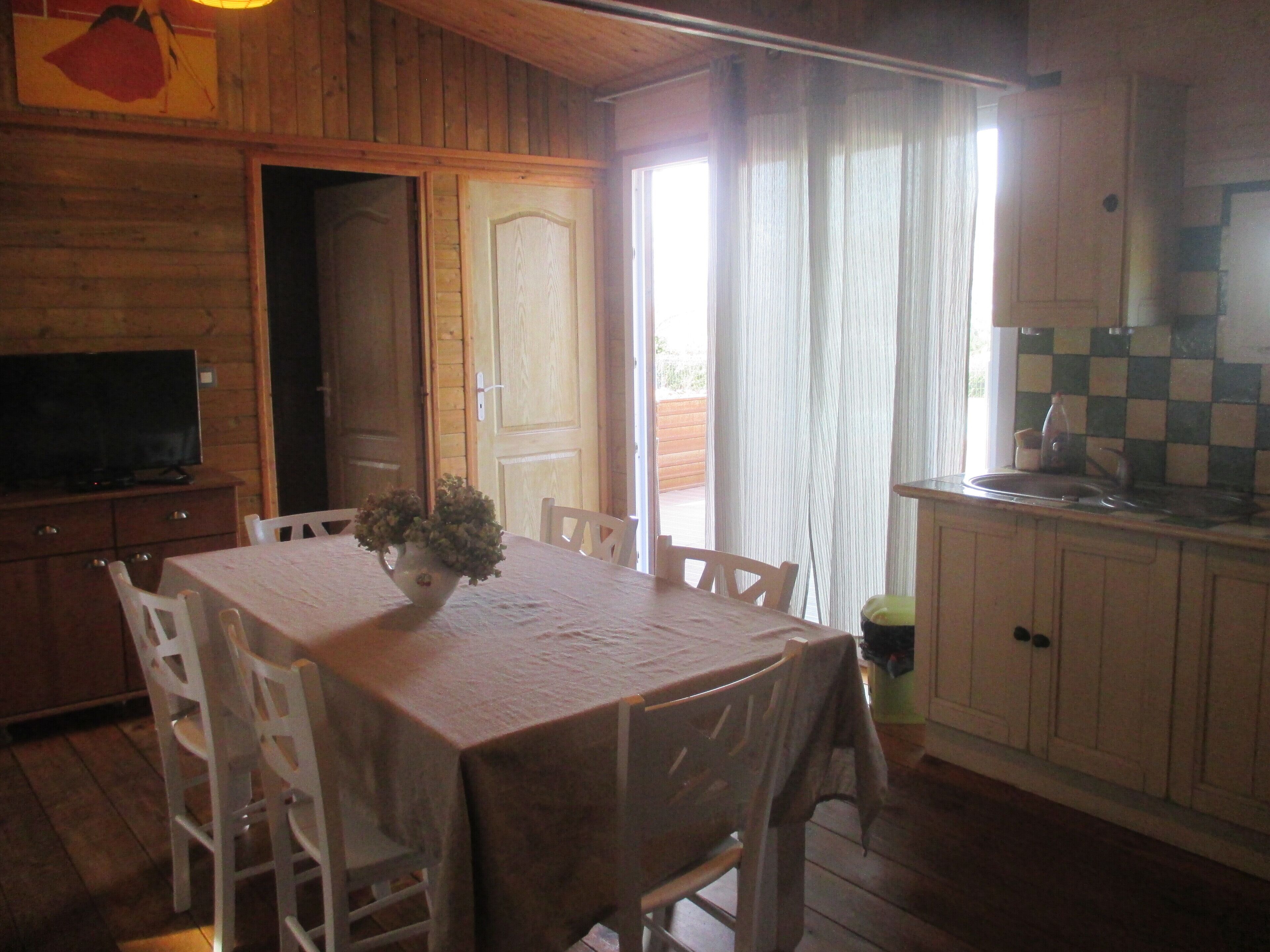 Un comedor acogedor con una mesa dispuesta para cuatro personas, un televisor sobre un mueble de madera y una zona de cocina con un fregadero y armarios.