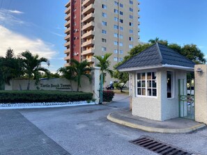A gated entrance to Turtle Beach Towers with a small building, palm trees, and a tall residential building in the background. - Beachfront Bliss | 1-BR condo with Panornomic sea-View (Ocho Rios)