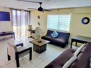 A living room with a sofa, coffee table, chairs, a TV, and a ceiling fan. - Beachfront Bliss 1-BR condo with Panoramic sea-View (Ocho Rios)