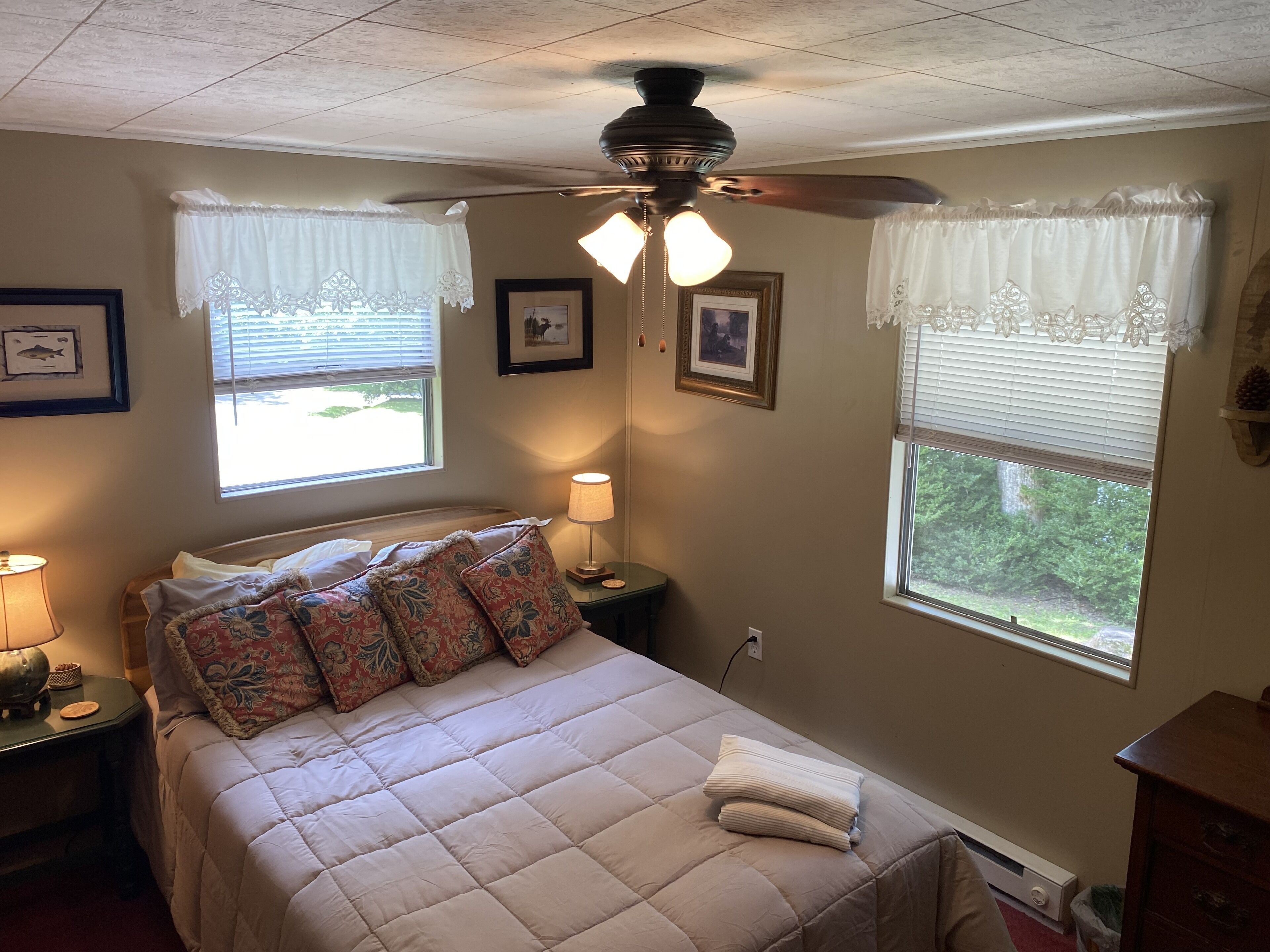 A bedroom with a bed, two bedside tables, a ceiling fan, and two windows with curtains.