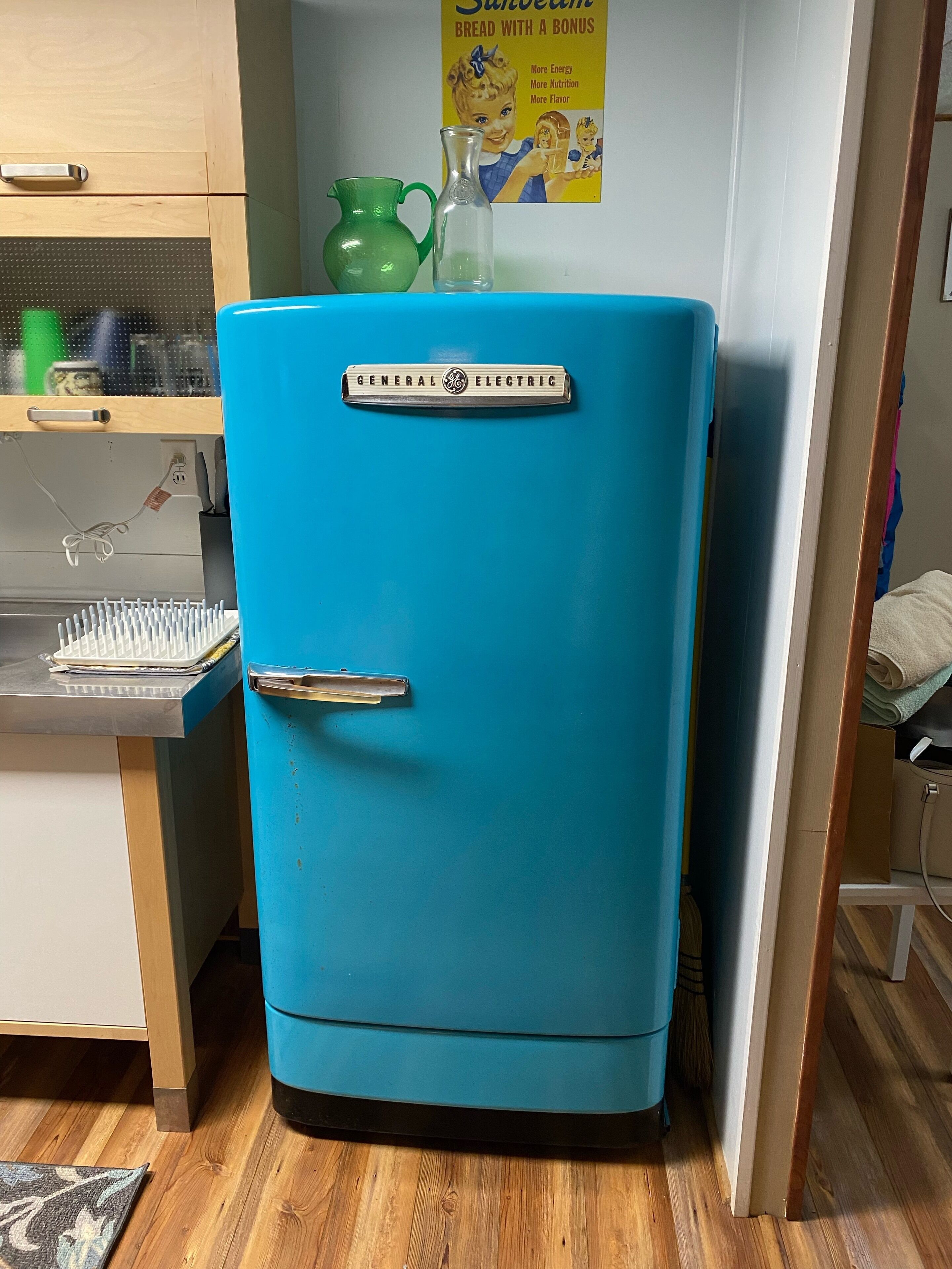 A teal-colored General Electric refrigerator in a kitchen with wooden cabinets, a green pitcher, and a vintage advertisement on the wall.