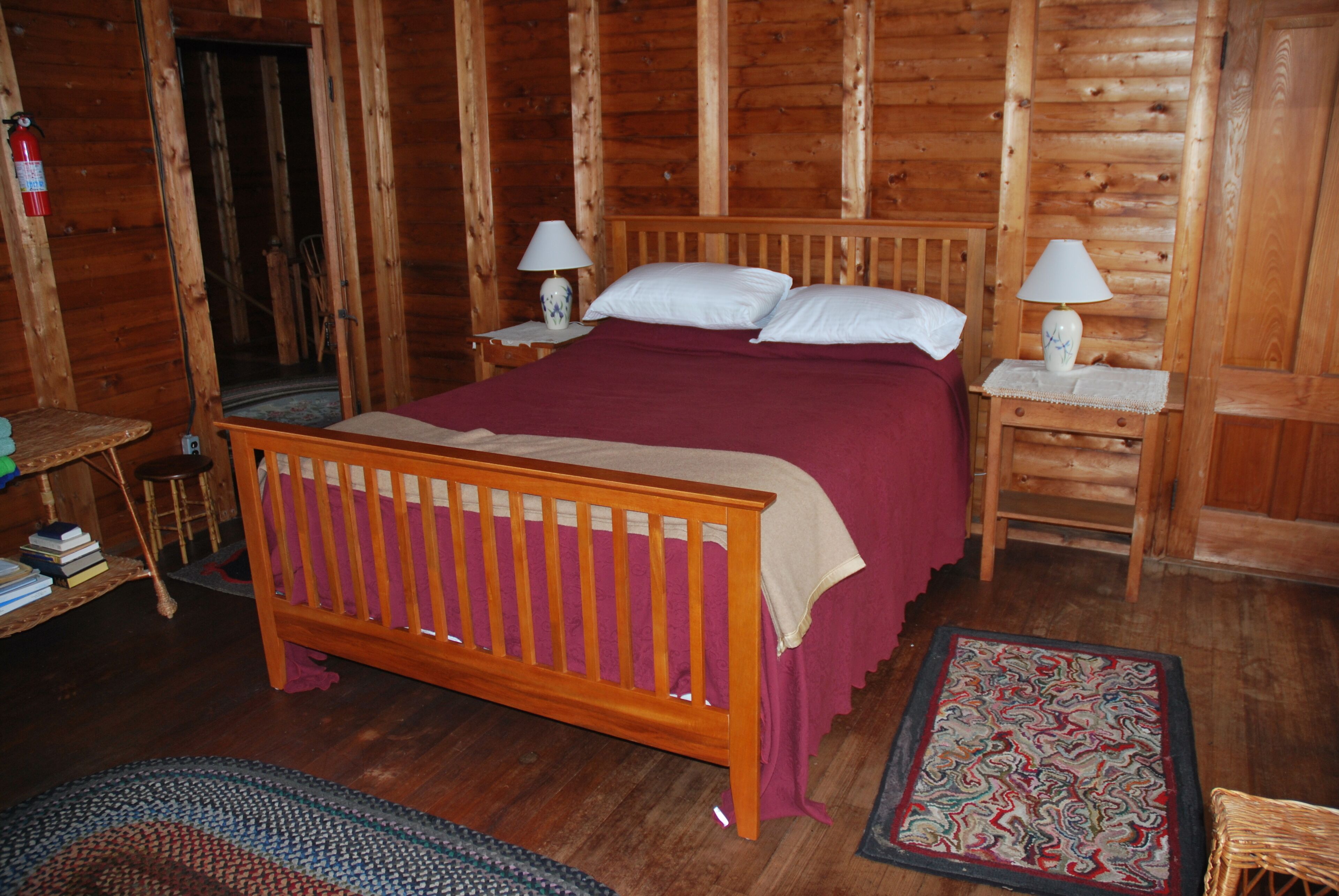 A wooden cabin bedroom with a wooden bed, two bedside tables with lamps, a small stool, a chair, and a patterned rug.