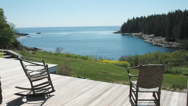 Une terrasse dotée de deux fauteuils à bascule, offrant une vue sur une mer calme et une côte bordée de forêts.
