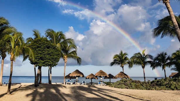 A beach with palm trees, beach chairs, and thatched umbrellas under a clear sky with a rainbow.