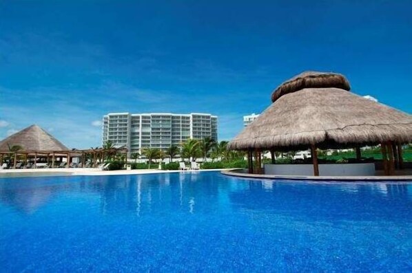 A resort pool with thatched umbrellas, a large building, and a clear blue sky.
