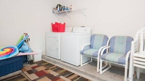 A laundry area with a washing machine, two chairs, a red basket, and a shelf with toiletries. - LAKE BERKLEY-(1059LB) - 4BR 3BA Pool Home, 2 Master Suites, gated Resort (Kissimmee)