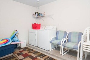A laundry area with a washing machine, two chairs, a red basket, and a shelf with toiletries.