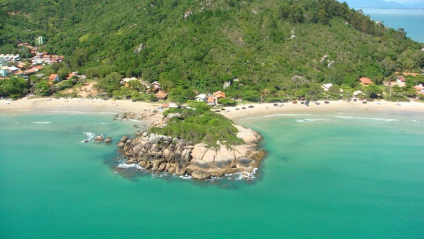 Vista aérea de una zona costera con una isla rocosa rodeada de aguas turquesas, una playa de arena y colinas verdes al fondo con edificios.