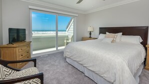 Een slaapkamer met een groot bed, een plafondventilator, een televisie op een houten kast en uitzicht op het strand door een glazen schuifdeur.