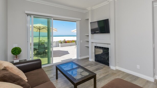 Een moderne woonkamer met een glazen tafel, een open haard, een aan de muur bevestigde televisie en een schuifpui naar een buiten terras met parasols.
