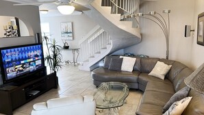 Un salon moderne comprenant un canapé d’angle, une table basse en verre, un téléviseur à écran plat, un escalier et un coin repas avec une table et des chaises en verre.