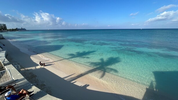 Une plage avec une eau turquoise et limpide, des palmiers qui projettent leur ombre et quelques personnes se détendant sur le sable.