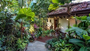 Un exuberante jardín con diversas plantas tropicales y una terraza de madera que conduce a un edificio con tejado de baldosas.