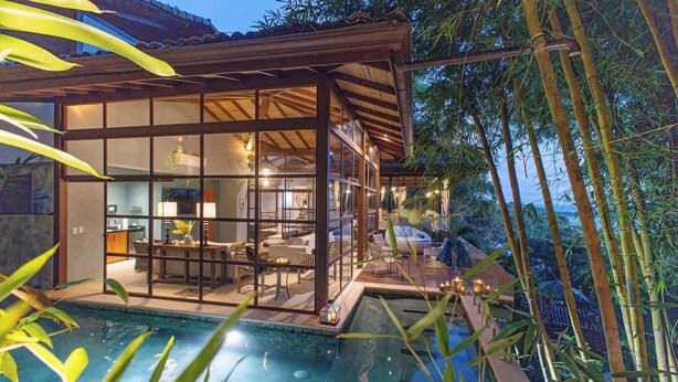 Una casa moderna con fachada de vidrio, piscina y árboles de bambú al fondo.