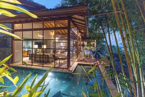 Una casa moderna con facciata in vetro, una piscina e alberi di bambù sullo sfondo.