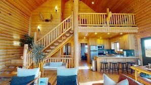 A log cabin interior with a central staircase, a kitchen area, and a dining space with bar stools. - Pearadise Peaks* Private Hot Tub, Wifi, 6Br, 3Ba (Island Park)