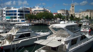 Un puerto deportivo con varios yates amarrados, una torre del reloj y un edificio moderno de fachada azul.