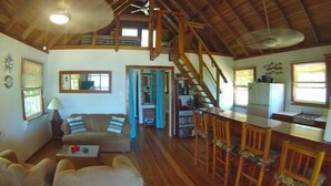 A spacious living area with a high ceiling, wooden beams, a sofa, a coffee table, and a dining area with chairs and a kitchen counter.