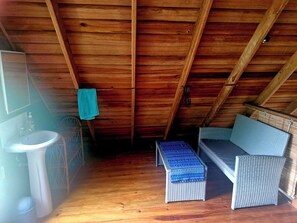 A small, enclosed attic room with a wooden ceiling, a white sink, a wicker bench, and a blue patterned ottoman.