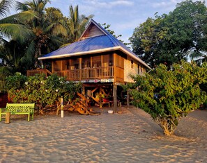 Una casa in legno con un balcone, affacciata su una spiaggia sabbiosa con palme sullo sfondo.