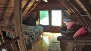 An attic bedroom with a sloped ceiling, wooden beams, a bed with striped bedding, a chair with cushions, a small table, and a window with blinds.