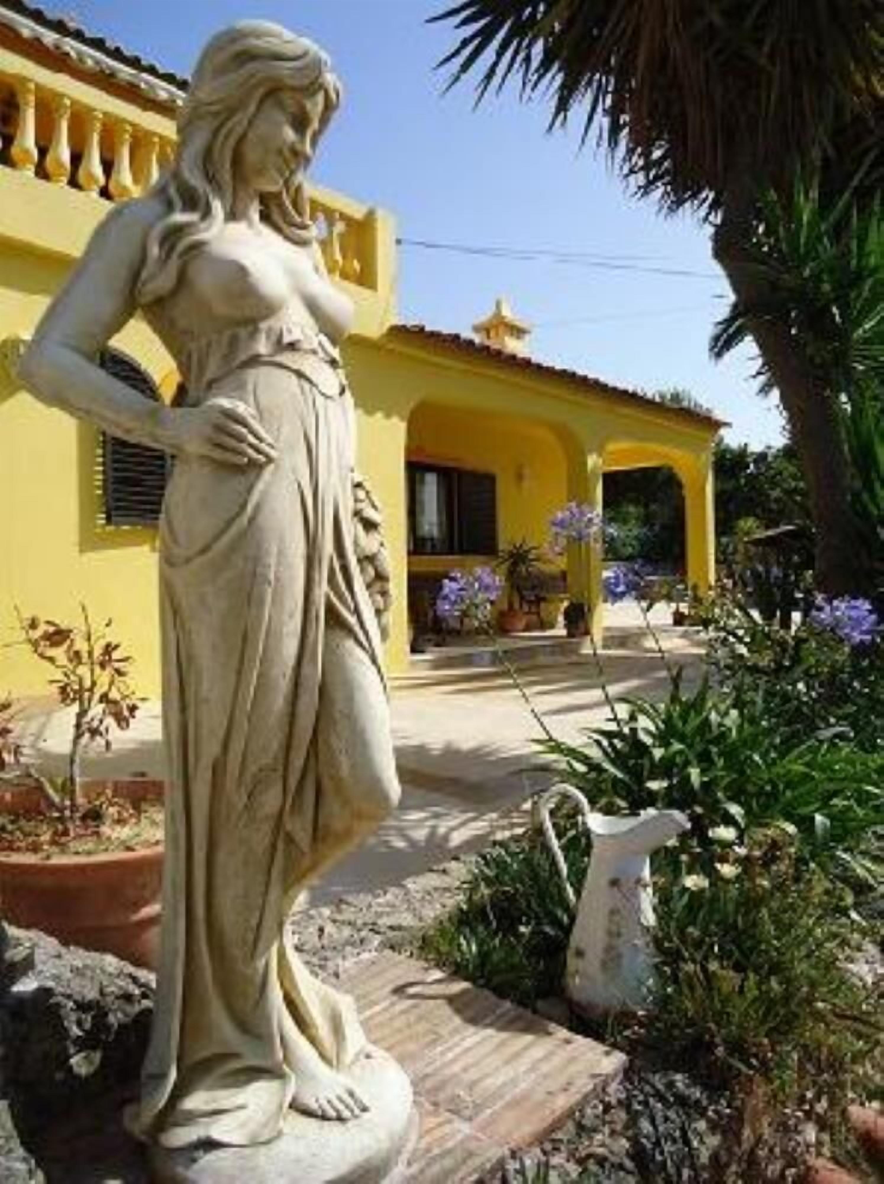 Una estatua de una mujer en un jardín, con un edificio amarillo al fondo.