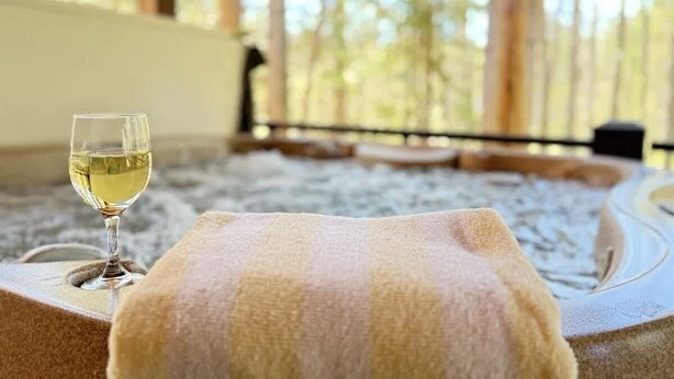 Un cadre de spa avec un verre de vin blanc posé sur une serviette, un bain à remous et une vue sur les arbres.