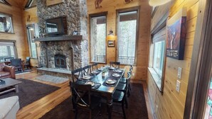 L’intérieur d’un chalet en bois est doté d’une cheminée en pierre, d’une table à manger dressée pour six personnes et de grandes fenêtres.