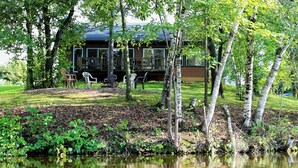 Una cabaña junto al lago con un balcón, rodeada de abedules y un área verde con mobiliario exterior.