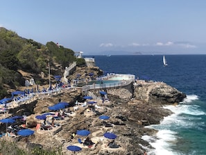 A rocky coastal area with a pool and lounge chairs under blue umbrellas, overlooking the sea.